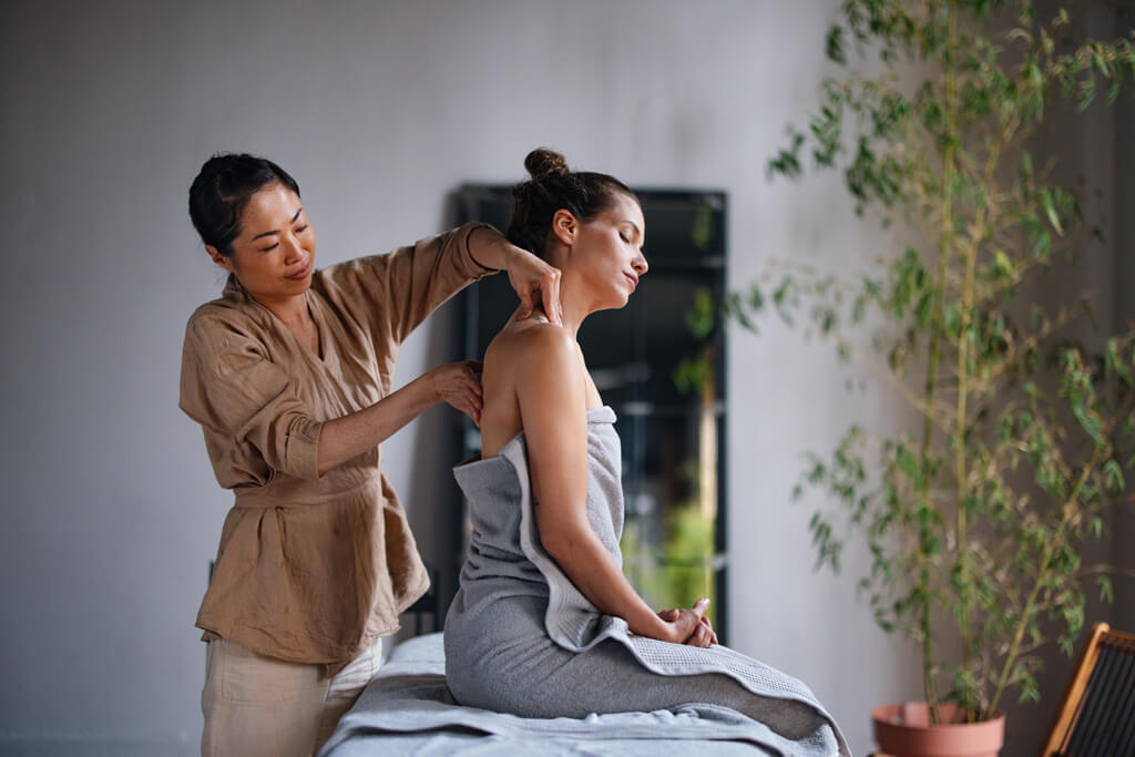 Schöne asiatische Therapeutin, die einer Frau, die auf dem Bett sitzt, eine Massage gibt